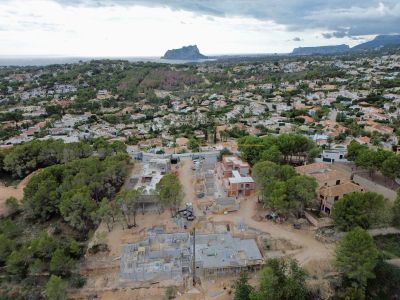 Vue aérienne d'un chantier de construction de villas avec fondations en béton et murs partiellement construits, dans une zone résidentielle avec vues lointaines sur mer et montagne.