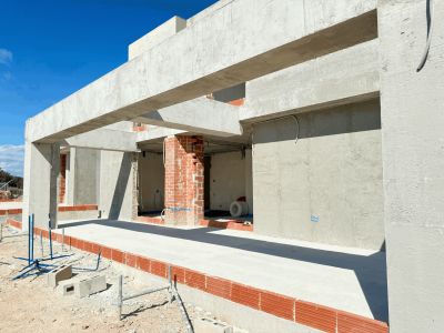 Structure en béton de la villa en construction, avec poutres, piliers et murs de briques apparentes sous un ciel bleu clair.