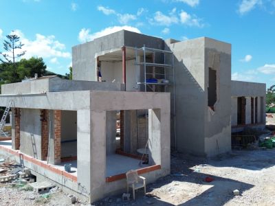 Structure en béton d'une villa moderne en construction, avec de grandes ouvertures pour les fenêtres et des échafaudages. Le chantier présente des matériaux de construction sous un ciel bleu.