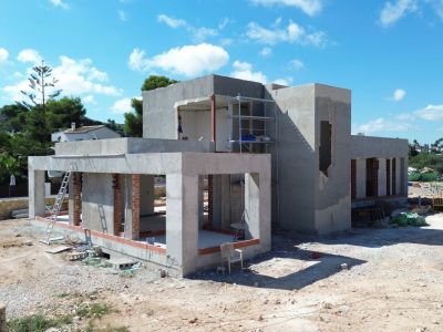 Villa en construction avec structure en béton et briques apparentes, échafaudages à l'étage supérieur et terrain de chantier environnant sous un ciel bleu dégagé.