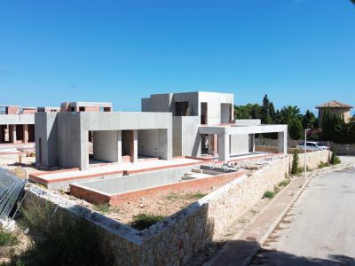 Villa moderne en construction, révélant sa structure en béton et l'excavation de la future piscine privée. Des murs en pierre délimitent la propriété le long de la rue résidentielle calme.