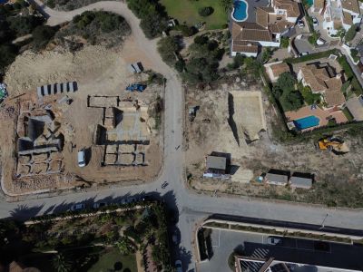 Vue aérienne de la phase initiale de construction d'une villa de luxe, révélant les fondations en béton et les parcelles excavées, au sein d'un quartier résidentiel avec piscines.