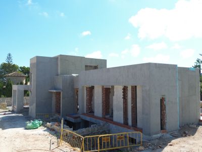 Vue extérieure de la villa en construction, présentant la façade enduite en gris, des piliers en briques apparentes et un terrain avec des barrières de chantier. Verdure et un bâtiment traditionnel en arrière-plan.