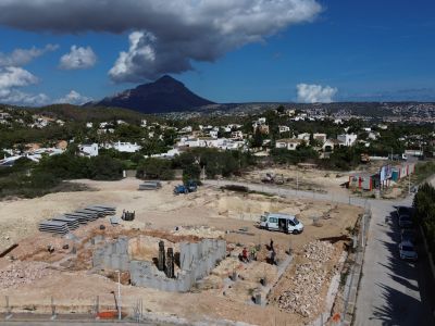 Vue aérienne du terrain à Jávea en construction, avec les fondations de la villa et des matériaux. Des maisons résidentielles et le Montgó sont visibles en arrière-plan.