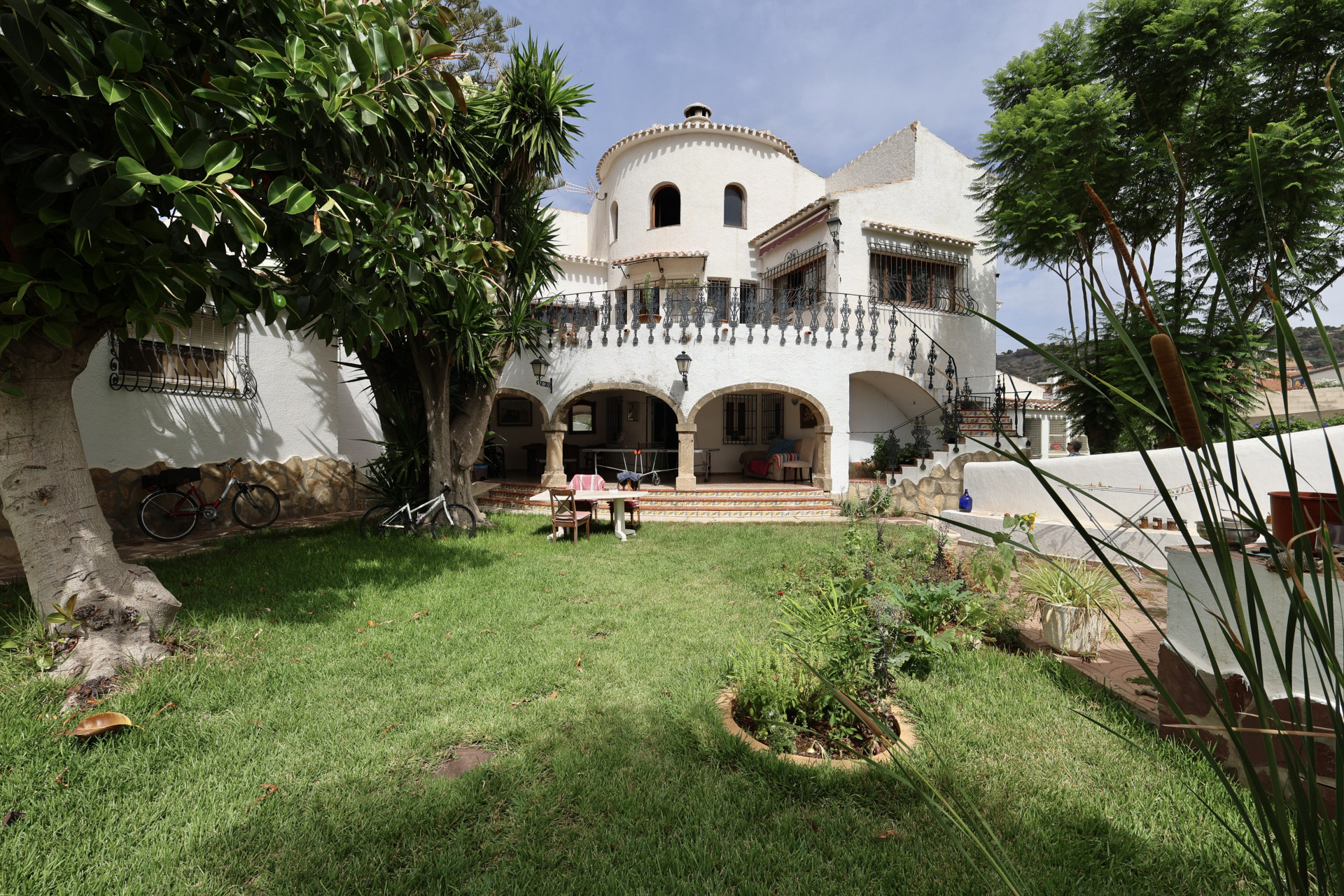 Vue sur le jardin avec pelouse, la façade blanche de la villa avec des arcades et une table à manger.