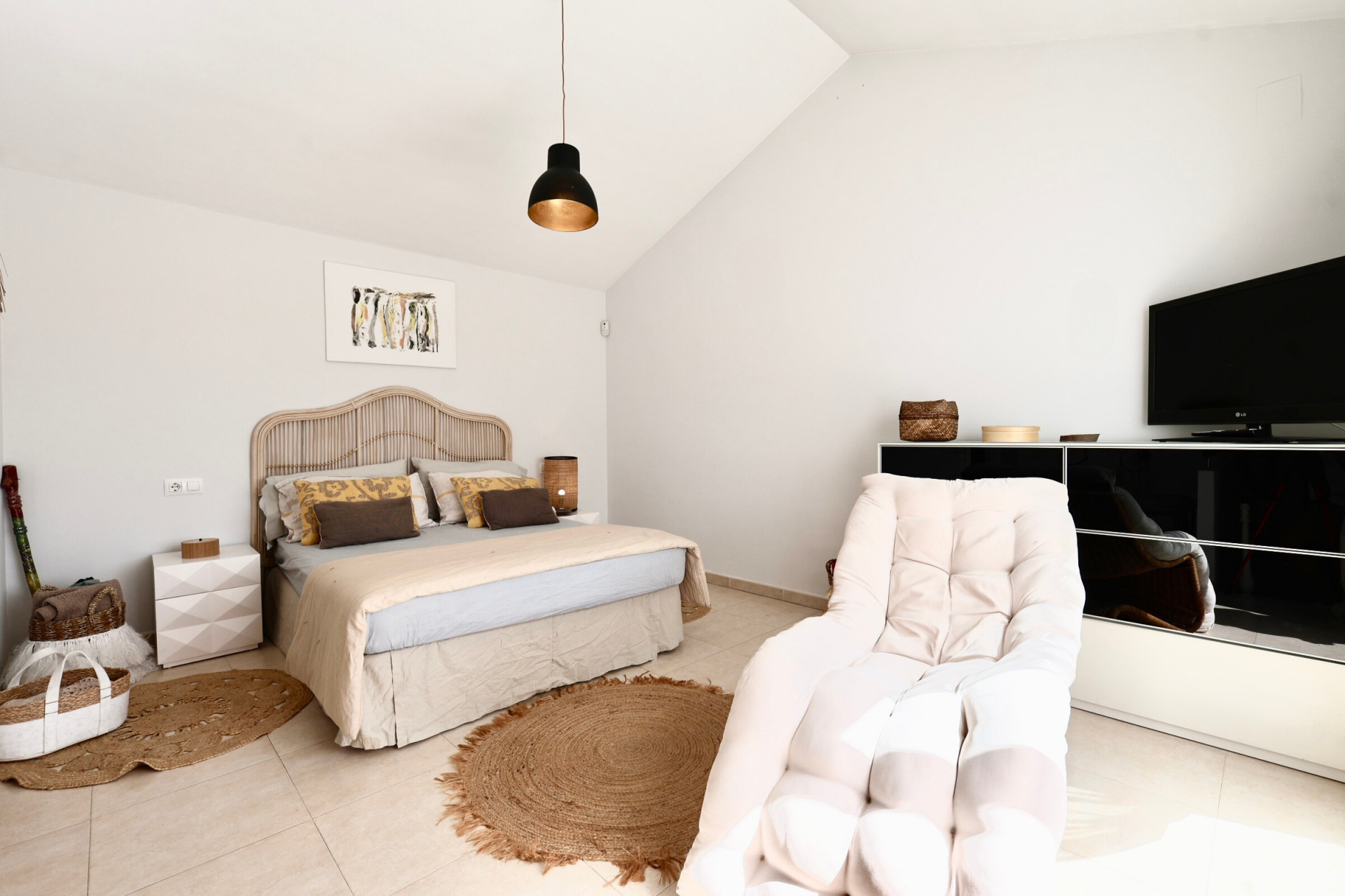 Chambre lumineuse avec lit en rotin, tapis en jute et fauteuil à côté d'un meuble TV noir.