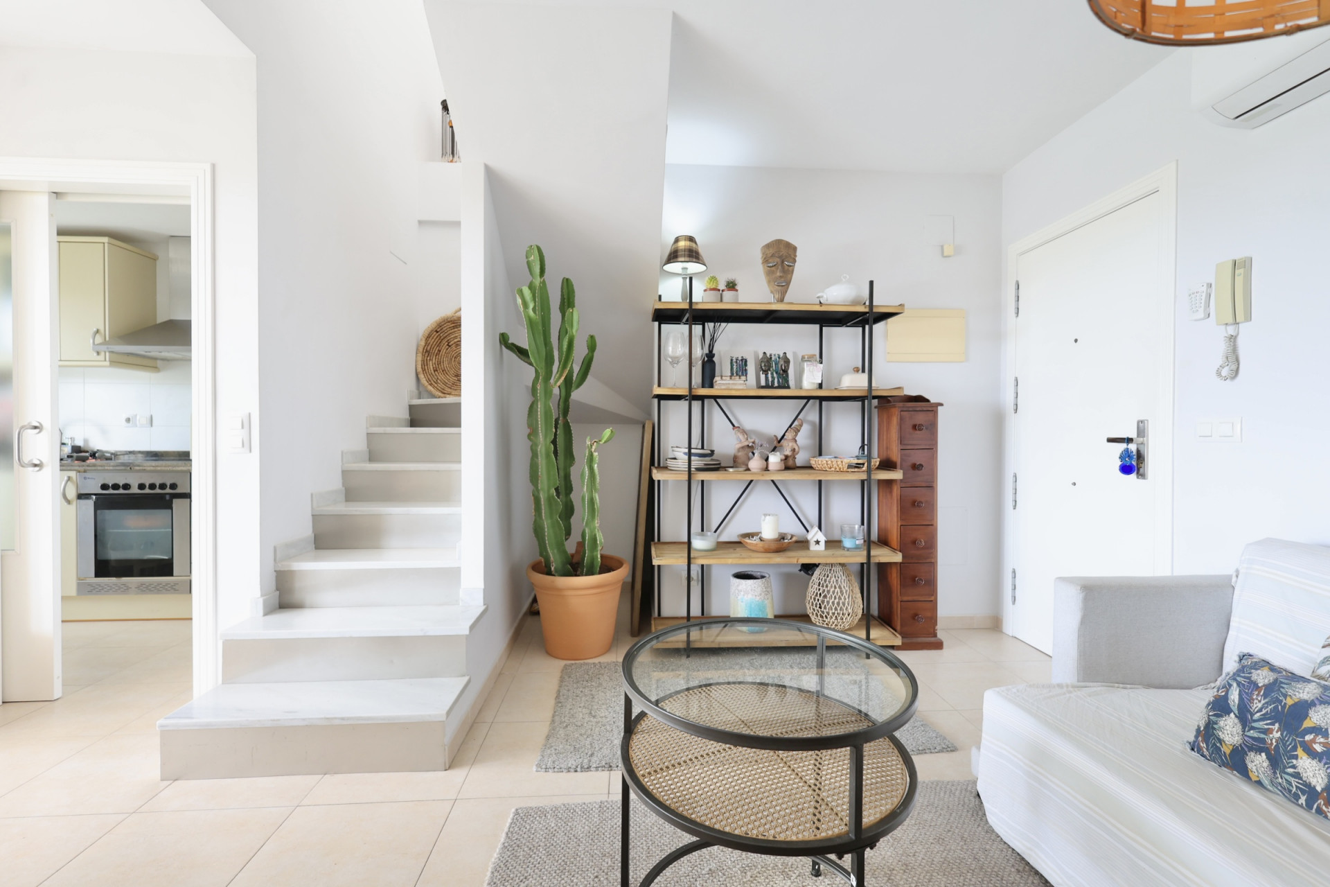Intérieur lumineux avec escalier blanc, étagère en bois et métal, et table basse en rotin.