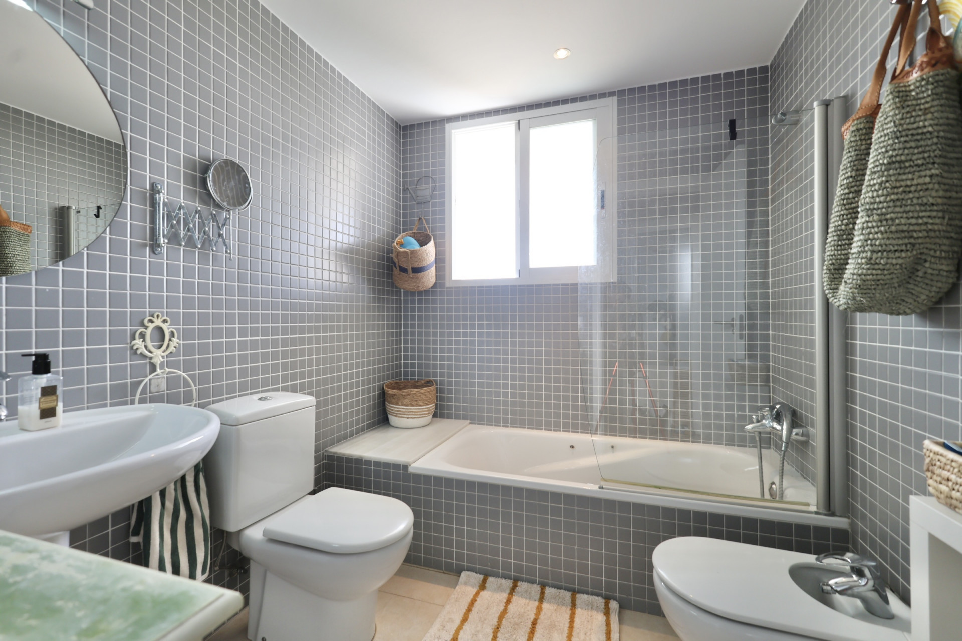 Salle de bain avec carrelage gris carré, lavabo sur pied, toilette et bidet. Une baignoire blanche est placée sous une fenêtre.