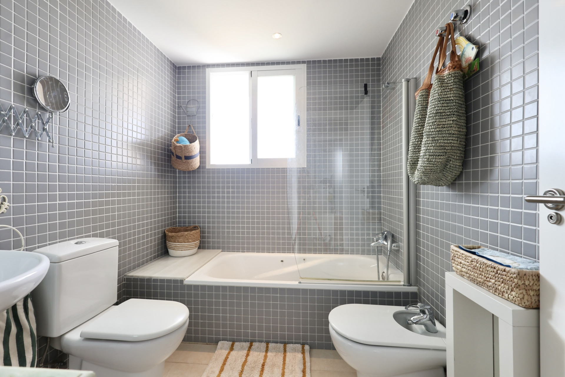 Salle de bain avec murs carrelés gris, baignoire avec pare-douche et éléments décoratifs en osier.