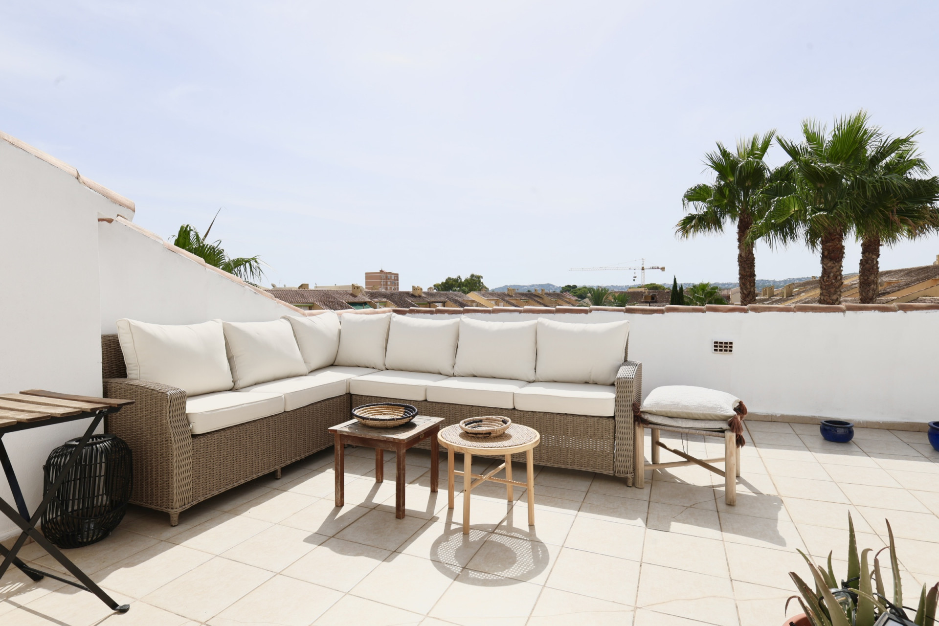 Terrasse avec canapé d'angle en osier et coussins blancs, donnant sur des palmiers et des toits.