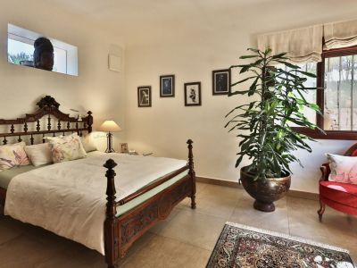 Chambre avec lit en bois sculpté, fauteuil en velours rouge et grande plante. Fenêtre à barreaux avec stores romains et fenêtre haute révélant une sculpture extérieure.