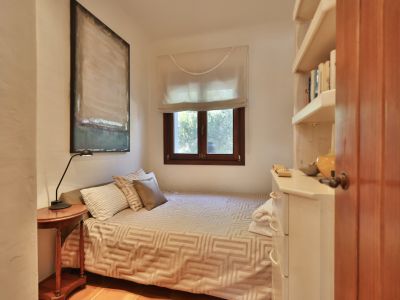 Chambre avec lit simple, table de chevet en bois et lampe. Fenêtre à cadre en bois avec store, étagère blanche intégrée avec livres et commode. Art abstrait et sol en terre cuite.