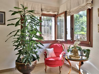 Coin salon intime avec fenêtres à cadres en bois offrant une vue sur le jardin, fauteuil rouge capitonné et grande plante en pot. Une table d'appoint présente une statue de Bouddha et une orchidée.