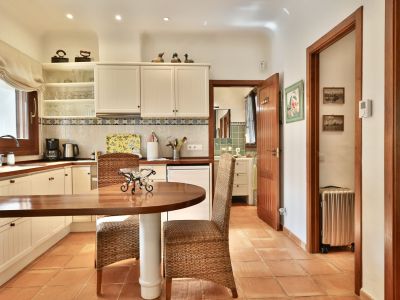 Cuisine fonctionnelle avec plans de travail en bois, armoires blanches et carrelage en terre cuite. Elle dispose d'une table ronde avec chaises en osier et d'un accès à une salle de bain.