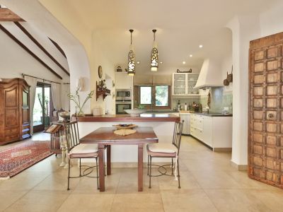 Cuisine ouverte avec comptoirs sombres, carrelage vert clair et appareils intégrés. Une table à manger en bois est placée près d'une porte décorative et d'un salon aux poutres apparentes.