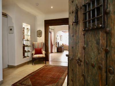 La porte d'entrée rustique en bois cloutée s'ouvre sur un couloir lumineux. On y voit un fauteuil rayé, une niche décorative avec étagères en verre et un tapis rouge sur carrelage clair.