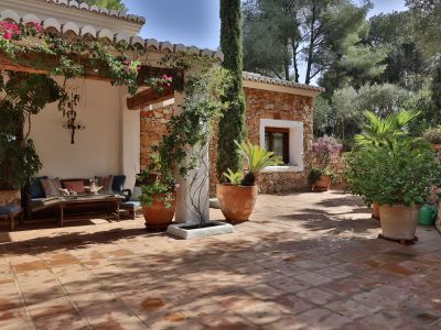Patio carrelé en terre cuite avec pergola ombragée de bougainvilliers, salon extérieur et annexe en pierre au milieu de plantes méditerranéennes et d'un cyprès.