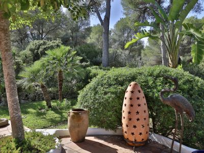 Patio de jardin avec sol en terre cuite, présentant un pot rustique, une lanterne étoilée et une statue de héron, encadré par une végétation méditerranéenne luxuriante et des palmiers.