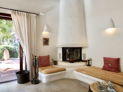 Salon chaleureux avec cheminée en stuc blanc et banquettes intégrées, s'ouvrant sur une terrasse carrelée et un jardin verdoyant.
