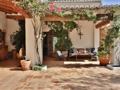 Terrasse couverte avec carrelage en terre cuite, poutres en bois apparentes et pergola ornée de bougainvilliers. Comprend un coin salon extérieur et un mur en pierre rustique.