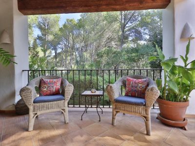 Terrasse couverte avec deux fauteuils en osier et coussins bleus, une table d'appoint en fer forgé, sol en terre cuite et vue sur la végétation luxuriante.