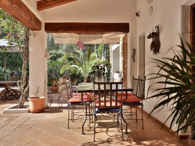 Terrasse couverte avec sol en terre cuite et poutres apparentes, meublée d'une table à manger en fer forgé, offrant une vue sur le jardin méditerranéen luxuriant.