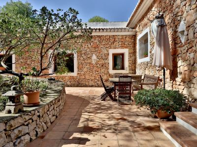 Terrasse en carreaux de terre cuite et murs en pierre naturelle, aménagée avec du mobilier de jardin en bois et un parasol. Des plantes en pot et un arbre apportent de l'ombre.