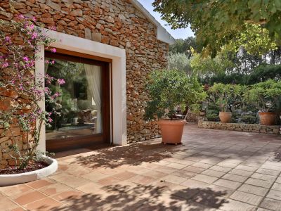 Terrasse extérieure dallée de terre cuite avec une façade en pierre sèche, encadrant une porte vitrée en bois. Une bougainvillée en fleurs et des plantes méditerranéennes ornent le jardin ensoleillé.