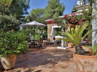 Terrasse extérieure en terre cuite, meublée d'un ensemble de repas en bois sous parasol, entourée de végétation méditerranéenne et d'une façade en pierre avec bougainvilliers.