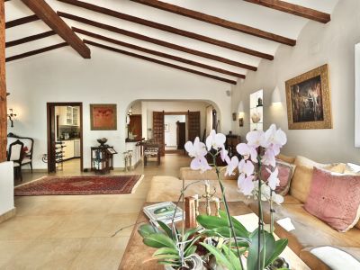 Vaste salon avec plafond voûté et poutres en bois apparentes, une colonne rustique, carrelage clair et un grand canapé sectionnel. Accès à la cuisine et autres pièces par des arches.