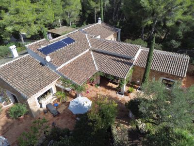 Vue aérienne de la villa avec toits en tuiles, murs en pierre et vaste terrasse en terre cuite. Panneaux solaires et pergola couverte sont visibles, entourés de végétation méditerranéenne.