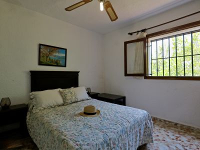 Chambre accueillante avec un lit double, linge de lit fleuri et tête de lit en bois foncé. Fenêtre à barreaux donnant sur la verdure extérieure et ventilateur de plafond.