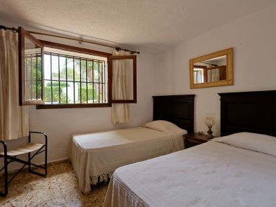 Chambre avec deux lits simples à têtes de lit en bois foncé, une table de chevet commune et une fenêtre grillagée donnant sur le jardin. Sol en terrazzo et miroir tressé.