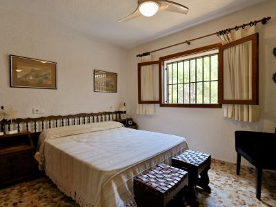 Chambre avec lit double, tête de lit en bois et carrelage à motifs. La fenêtre, dotée de volets et barreaux, offre lumière naturelle et vue sur la verdure.