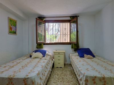 Chambre double avec deux lits simples, table de chevet, sol en terrazzo et fenêtre grillagée offrant une vue sur la verdure.