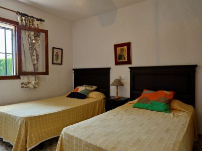 Chambre double avec têtes de lit en bois foncé, couvre-lits jaunes à motifs et coussins colorés. Fenêtre à barreaux et rideaux fleuris.
