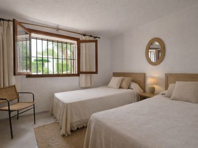 Chambre lumineuse avec deux lits simples, têtes de lit en bois clair et fenêtre ouverte avec barreaux de sécurité donnant sur un jardin verdoyant.