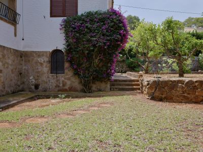 Coin extérieur avec base en pierre et mur blanc texturé, orné d'un bougainvillier violet. Fenêtre cintrée grillagée et escaliers en pierre vers le jardin.