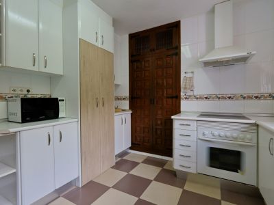 Cuisine équipée avec des armoires blanches, un four intégré et un micro-ondes. Une porte en bois foncé à panneaux et un sol à carreaux sont visibles.