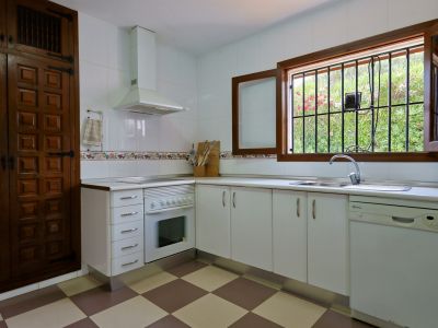 Cuisine fonctionnelle avec armoires blanches, sol carrelé à damier et fenêtre en bois grillagée donnant sur le jardin. Évier en acier inoxydable et lave-vaisselle intégrés.