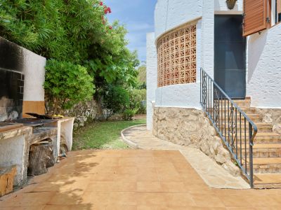 Espace barbecue extérieur avec terrasse carrelée, escalier avec rampe en fer forgé et façade en stuc blanc ornée d'un écran décoratif, entouré de verdure méditerranéenne.