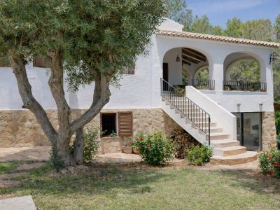 Extérieur de villa avec base en pierre et murs blancs, agrémenté d'un olivier mature. Un escalier extérieur mène à une terrasse couverte arquée avec balustrade en fer.