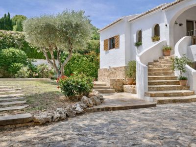 Façade blanche de la villa avec escalier en pierre courbé menant à l'entrée. Le jardin méditerranéen présente un olivier, des arbustes et des dalles au sol.