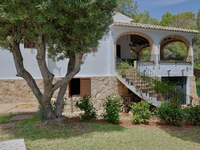 L'extérieur de la villa présente une façade blanche texturée sur une base en pierre, avec une terrasse couverte à arcades et un escalier extérieur carrelé, près d'un olivier.