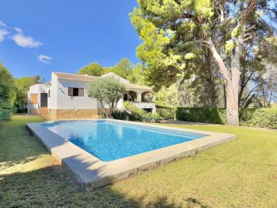 Piscine rectangulaire carrelée de bleu, entourée d'une pelouse verte soignée, avec une villa blanche à terrasse couverte et des arbres méditerranéens en arrière-plan.
