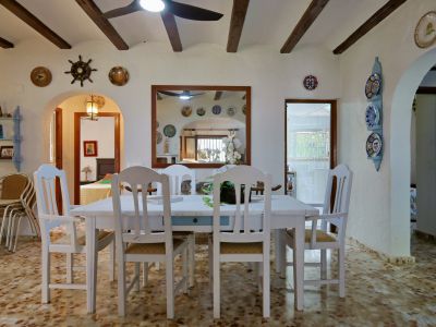 Salle à manger avec table et chaises en bois blanc, sol carrelé à motifs et plafond à poutres apparentes. Un grand miroir encadré reflète la pièce et sa décoration.