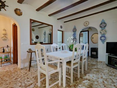 Salle à manger avec table et chaises en bois blanc, sol en terrazzo et plafond à poutres apparentes. Des arches et miroirs décoratifs structurent la pièce.