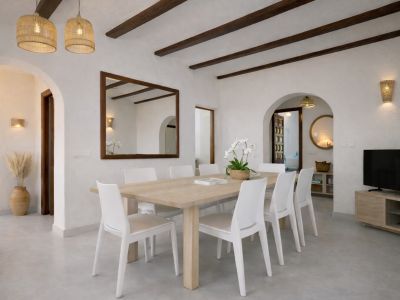 Salle à manger lumineuse avec table en bois clair pour huit et chaises blanches. Le plafond présente des poutres sombres, des suspensions en osier et un grand miroir encadré.