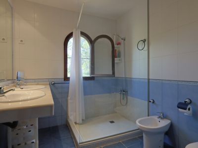 Salle de bain avec double vasque et plan de travail clair, murs carrelés blancs et bleus, et douche à l'italienne vitrée. Deux fenêtres en arc en bois apportent de la lumière naturelle.