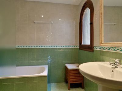 Salle de bain avec murs carrelés verts et beiges, baignoire à écran dépoli et lavabo sur pied. Fenêtre cintrée en bois avec verre texturé apporte lumière naturelle.